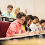 education, high school, university, learning and people concept - group of international students with notebooks writing in lecture hall