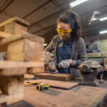 Female carpenter working with sander while standing surrounded with wood.