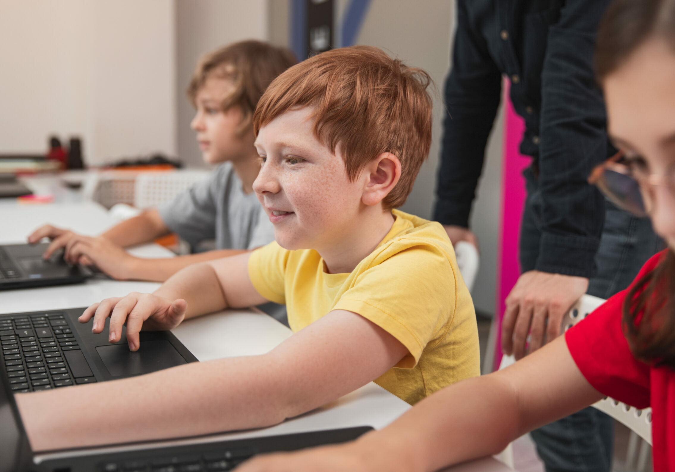 Positive smiling preteen schoolboy using laptop while studying computer programming with classmates during lesson in modern school